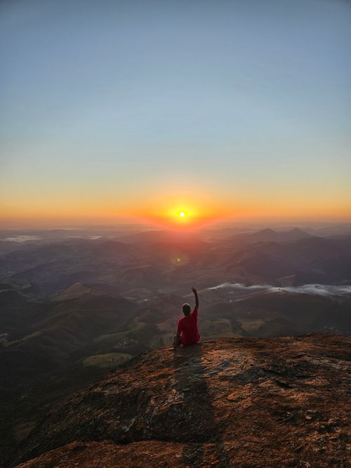 Free Person sitting on rock watches sunset over Aiuruoca mountains in Brazil, serene scene. Stock Photo
