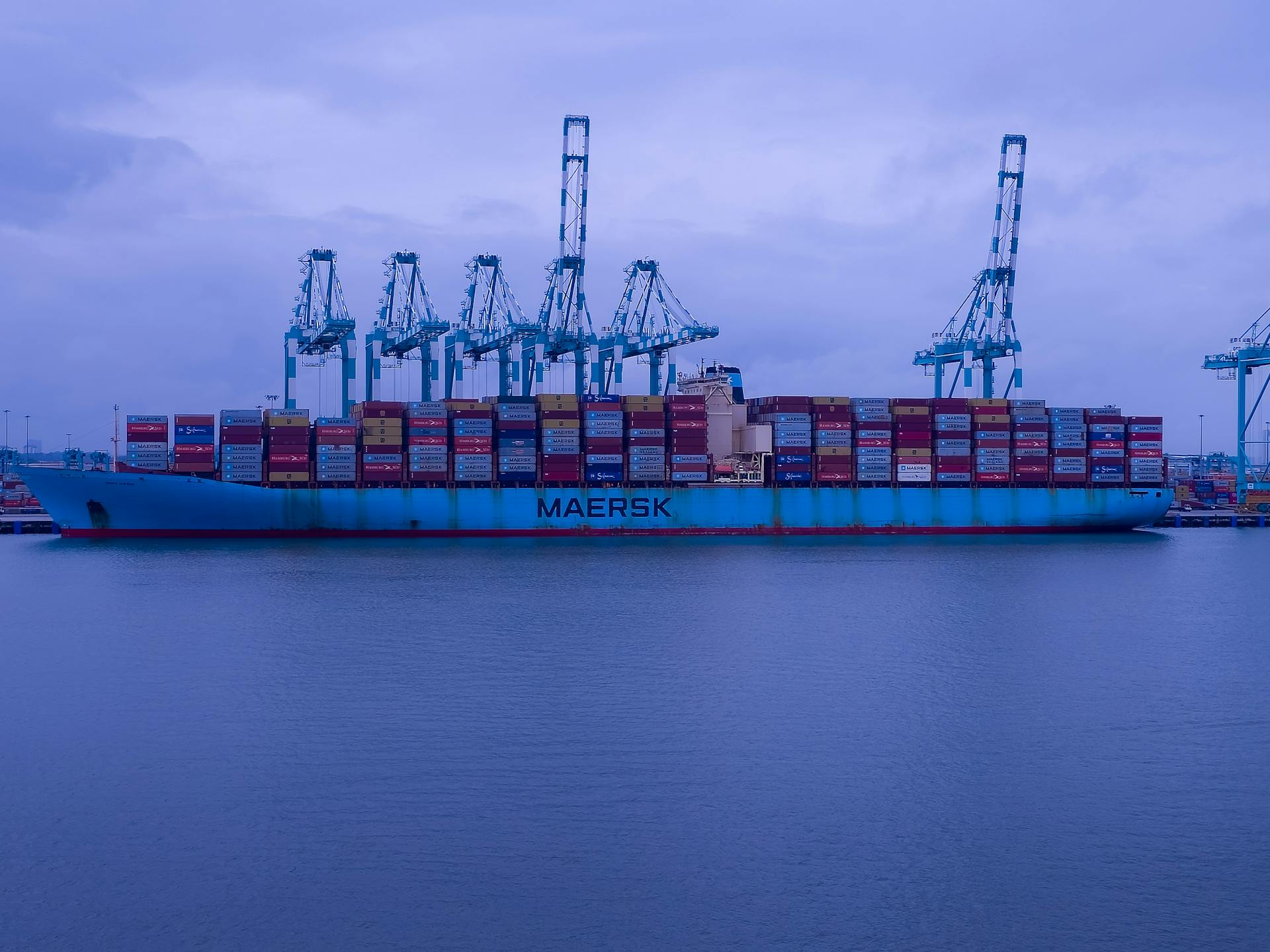 A Maersk cargo ship loaded with containers at an industrial port under a cloudy sky.