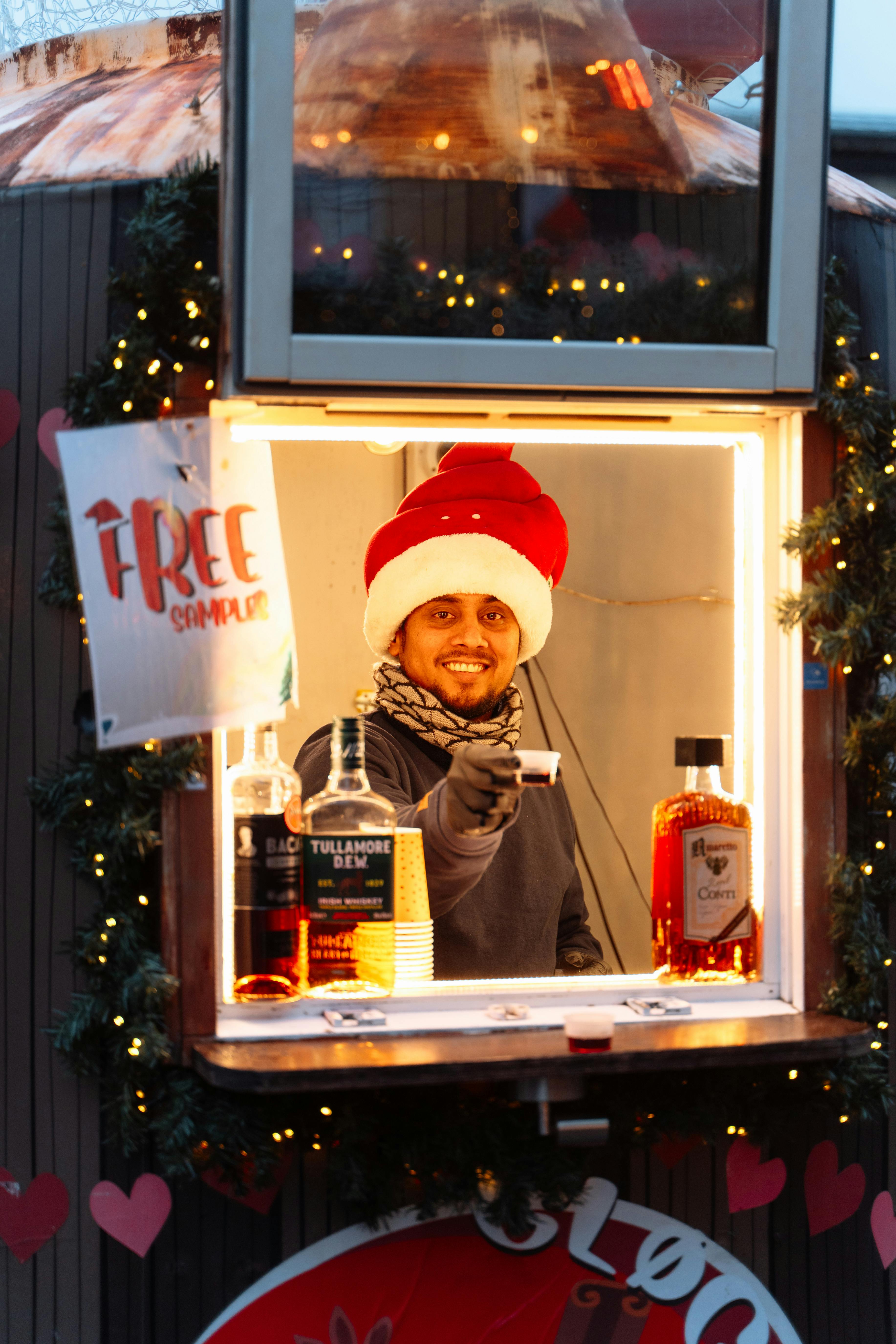 A joyful bartender in Santa hat offers drinks at a Copenhagen stand, decorated with lights.