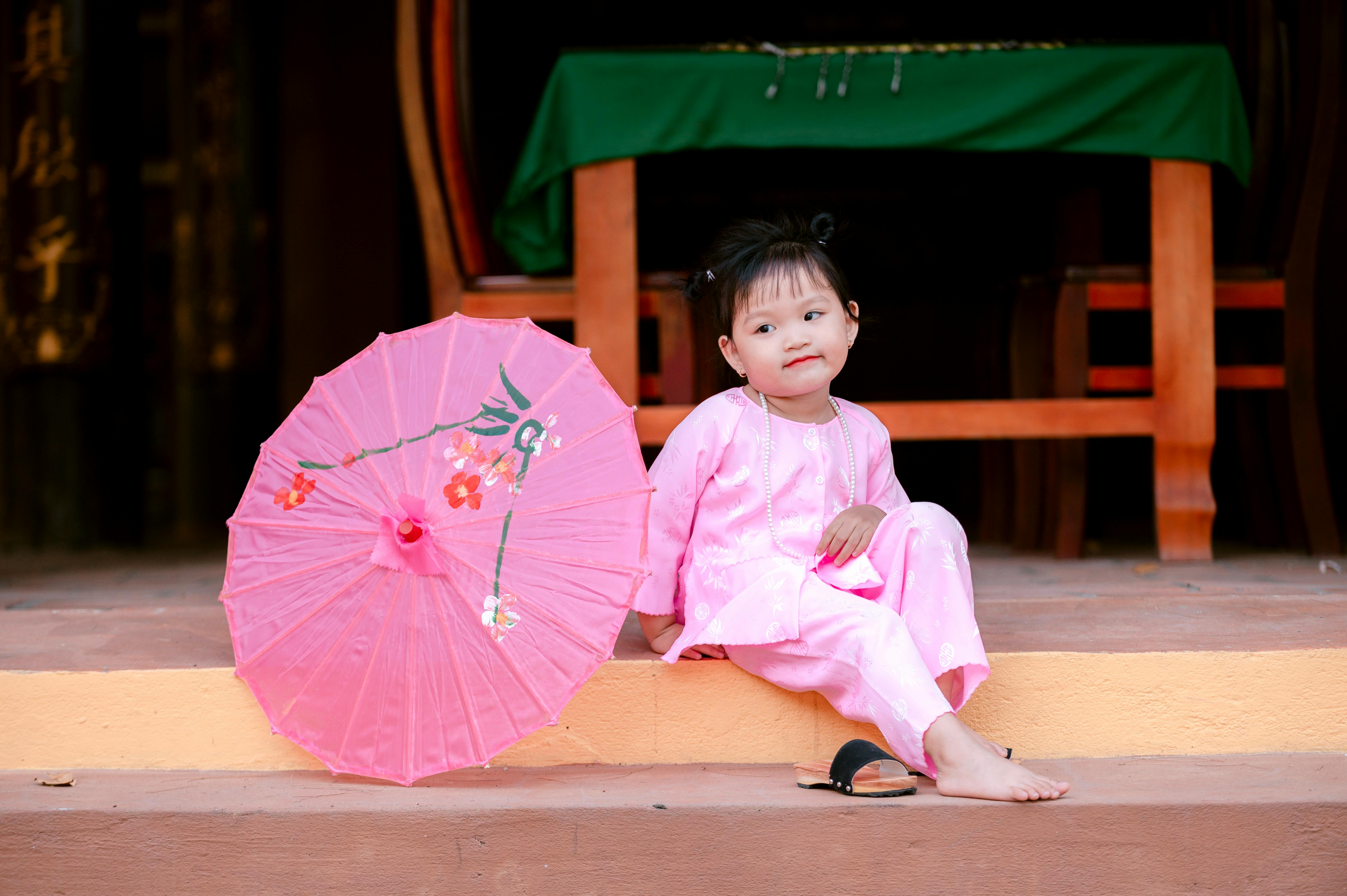 Child wearing pink traditional attire next to a decorated pink umbrella, sitting outdoors.