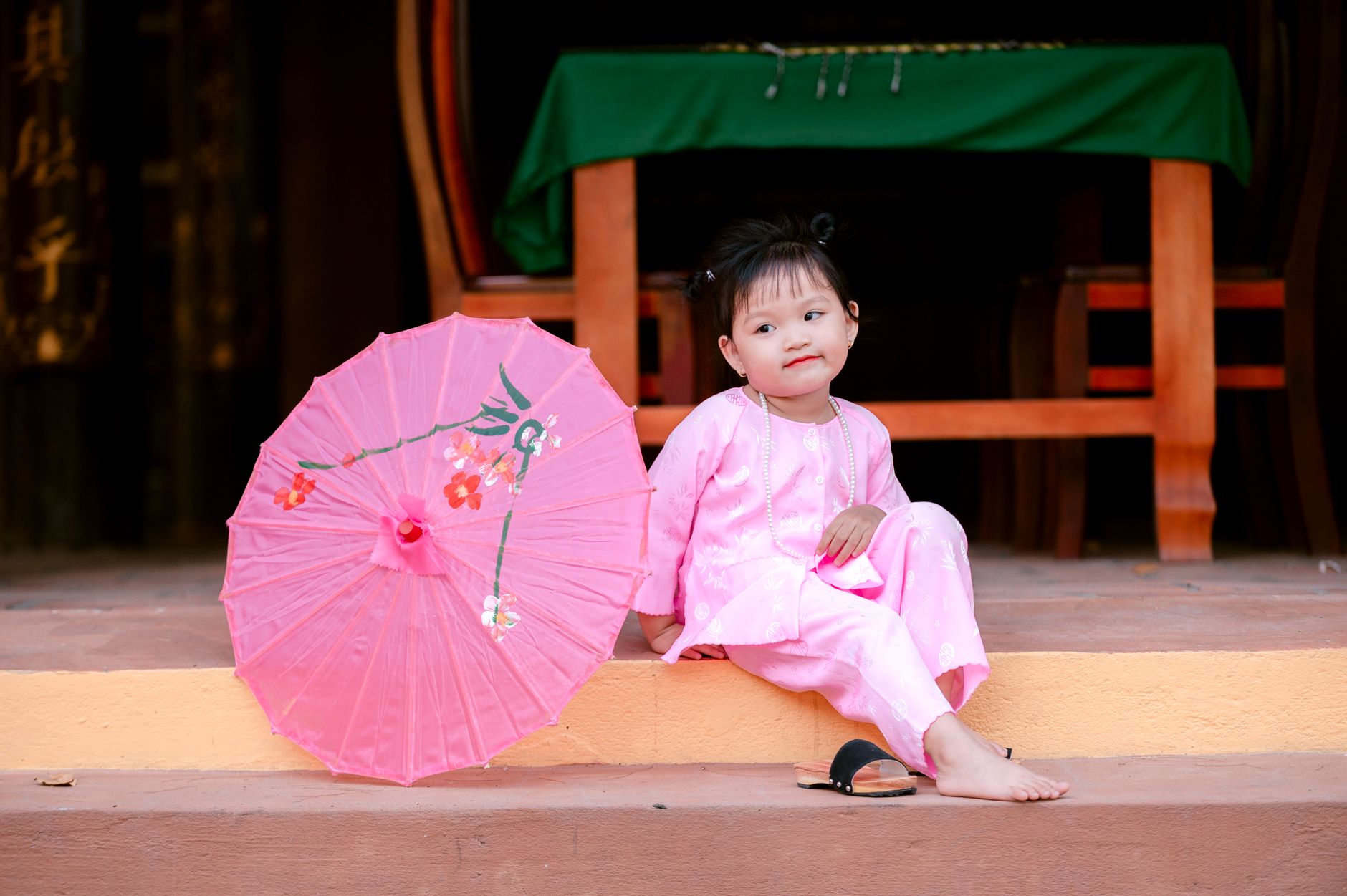 Child wearing pink traditional attire next to a decorated pink umbrella, sitting outdoors.