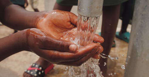 Close-up Of A Child's Hands Catching Water From The Spout Of A Water Pump