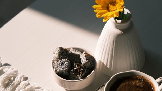 A cozy breakfast setup with coffee, sweets, and a flower in warm sunlight.