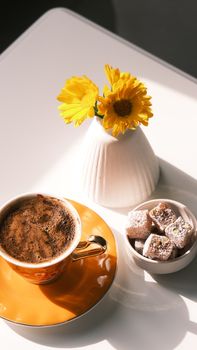 Sunlit coffee set with yellow flowers and confections on a bright table.