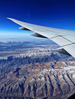 View from airplane window showing vast snowy mountain range under a clear blue sky.