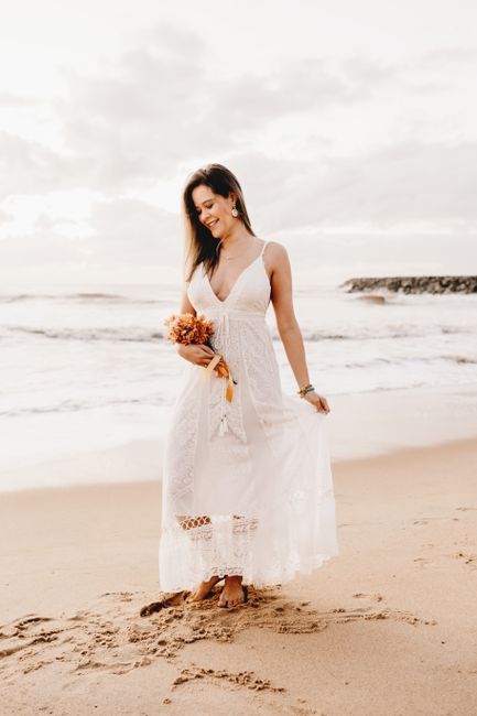 A woman in a white wedding dress stands barefoot on a sandy beach, holding a bouquet, with waves in the background.