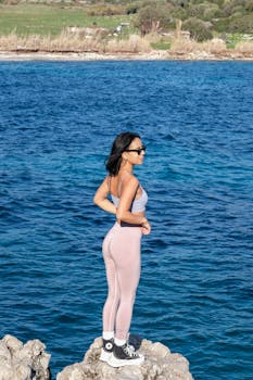 Woman in activewear stands on rocky shore, enjoying a scenic coastal view.