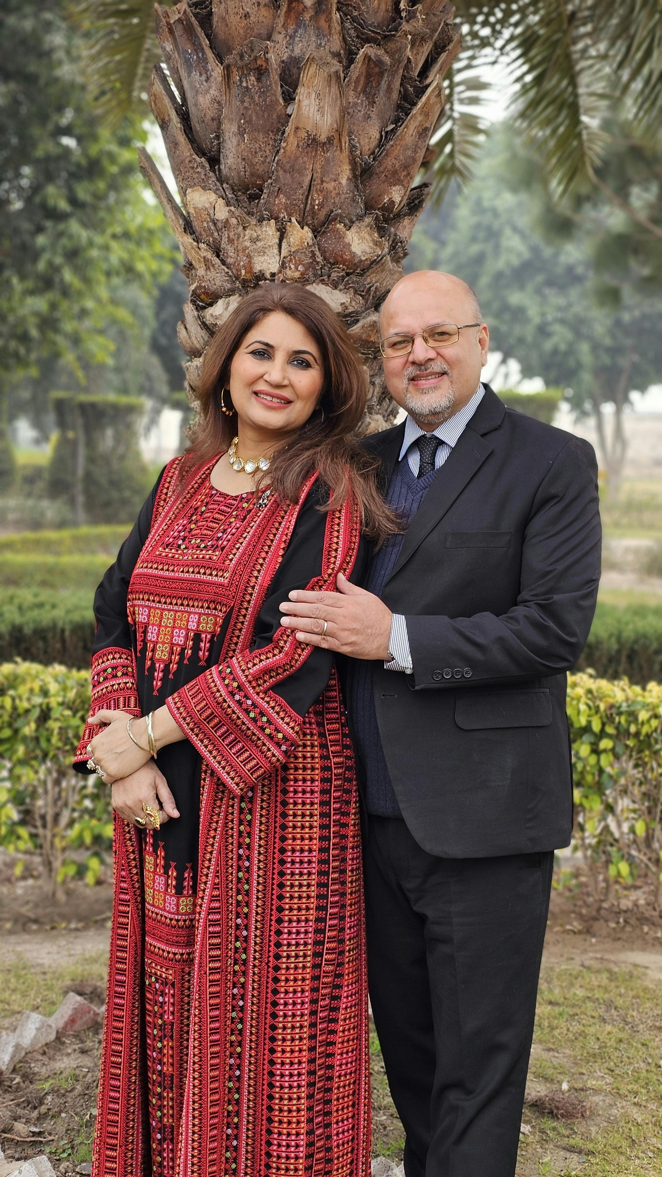 Stylish couple dressed formally standing outdoors beside a palm tree, exuding warmth and confidence.