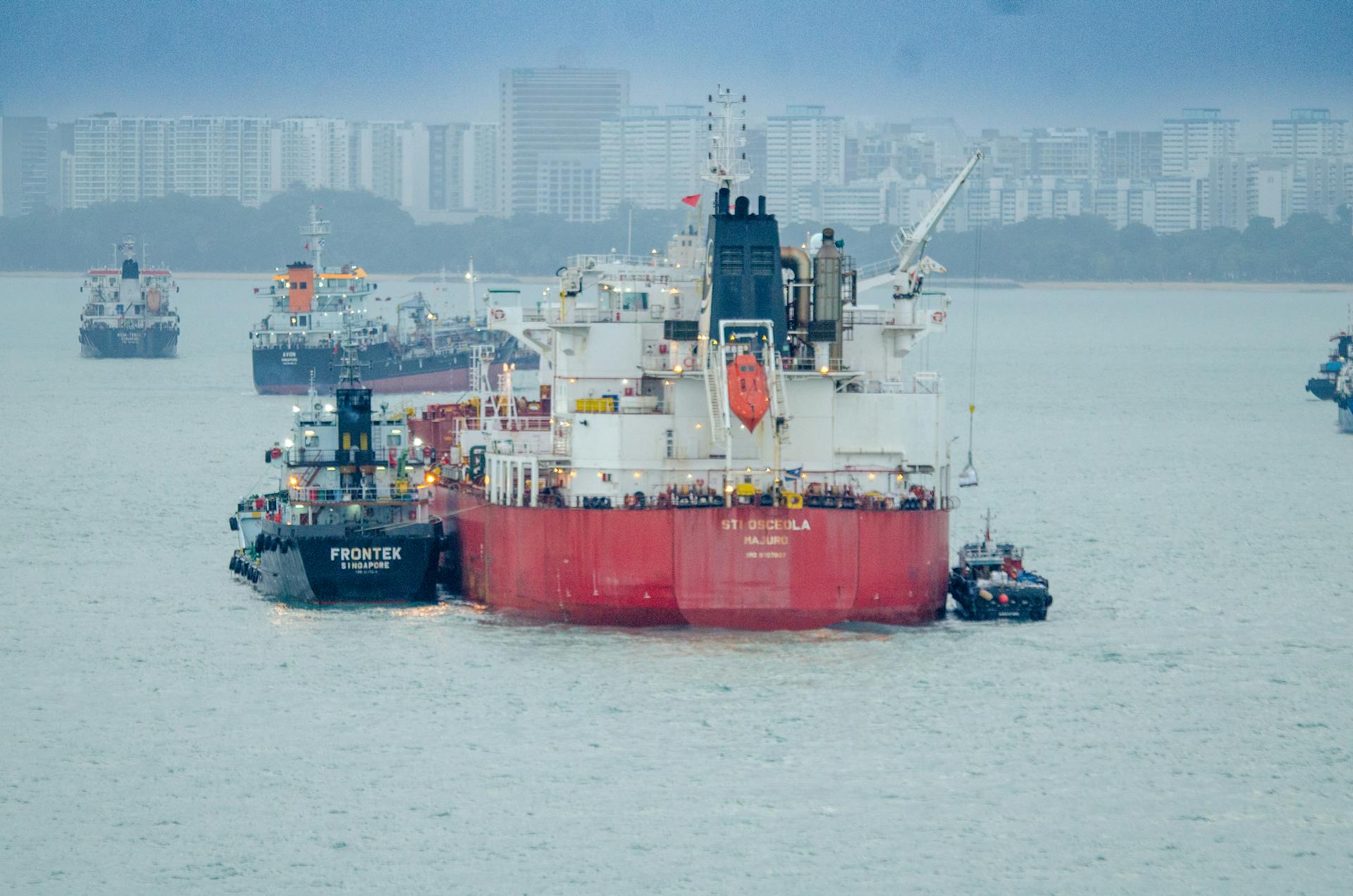 Cargo ships and tugboats navigating a bustling urban harbor against a city skyline backdrop.