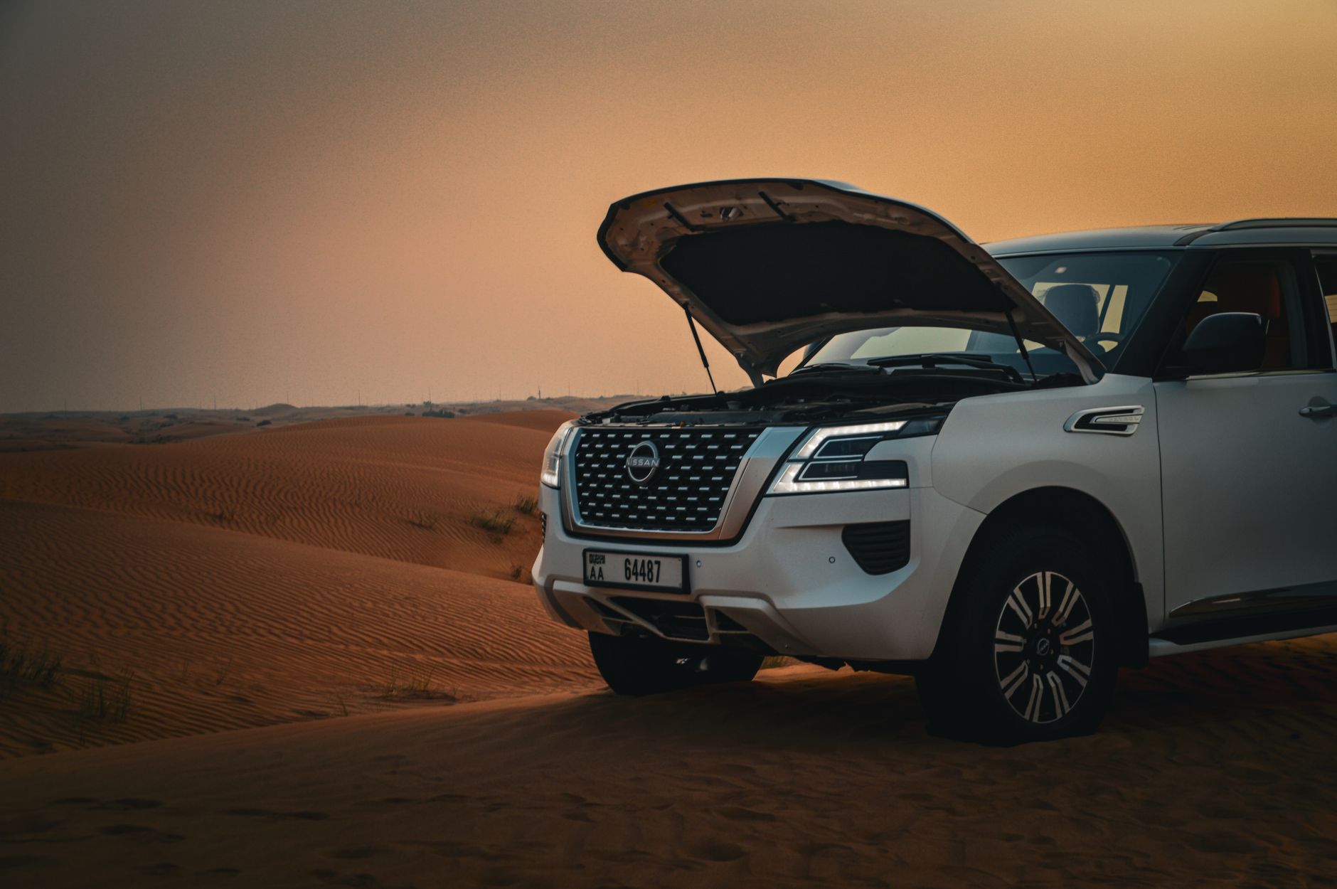 A white SUV with an open hood parked on scenic desert dunes during sunset.