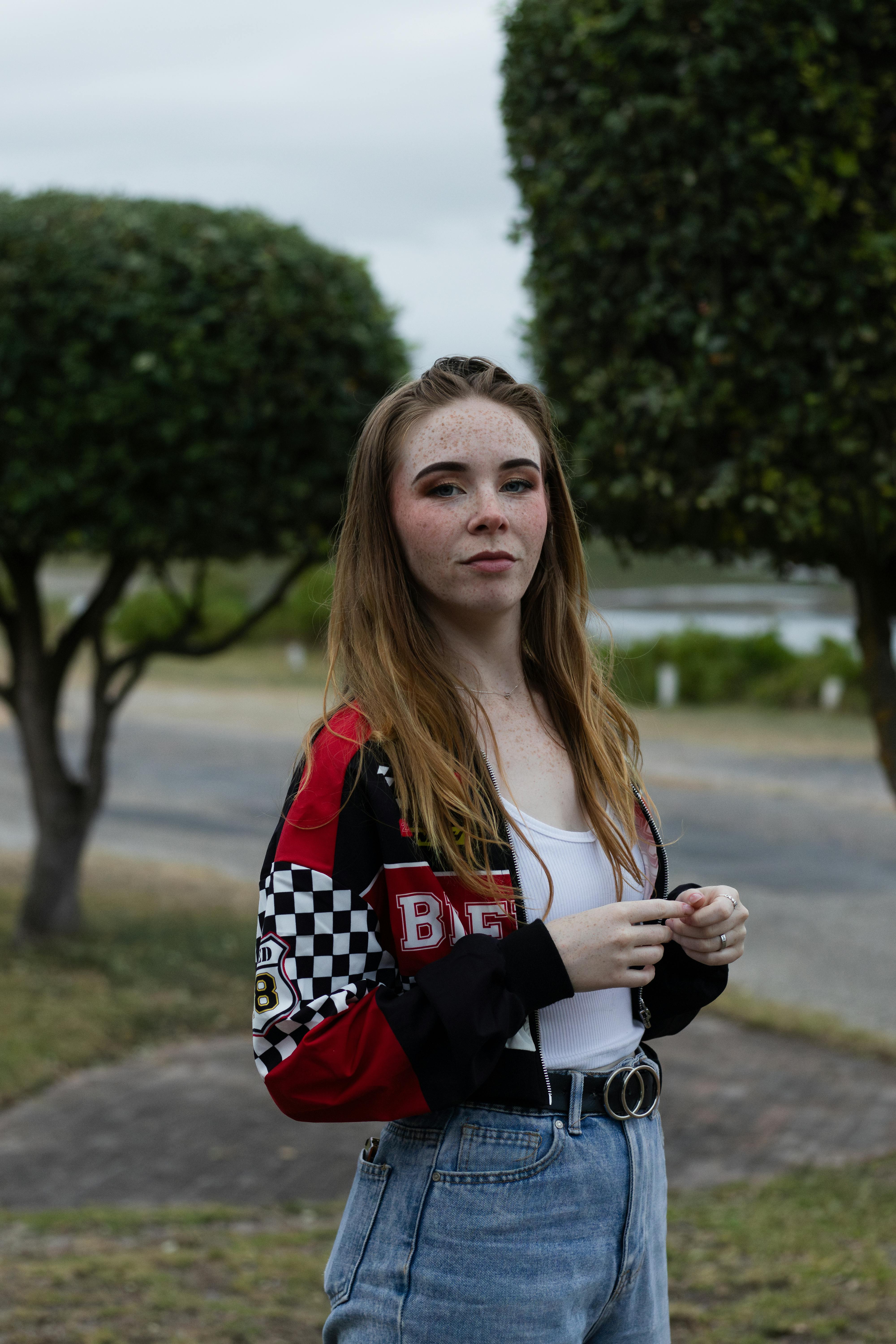 Young woman in casual attire poses outdoors with tree background, exuding confidence.