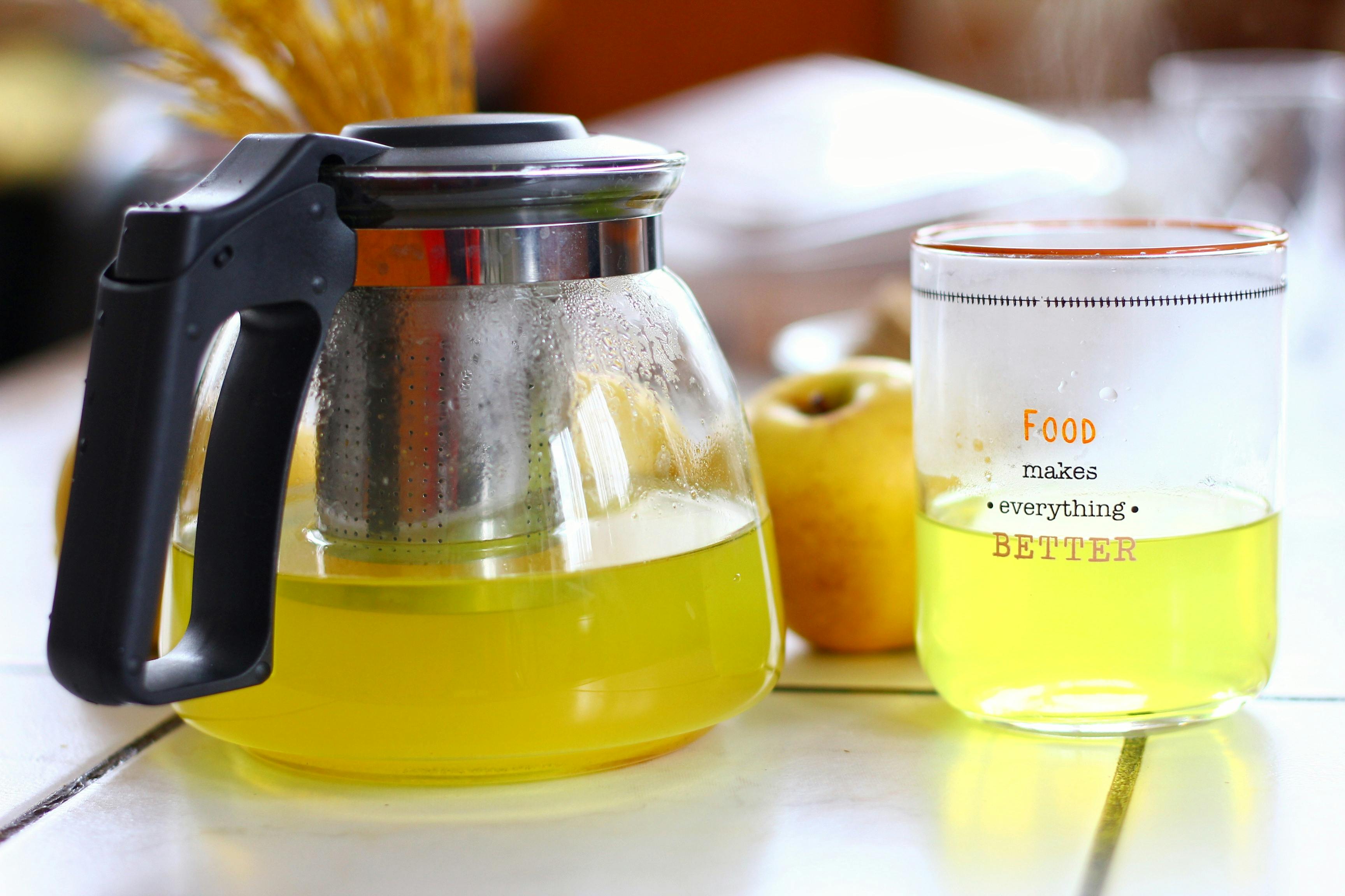 A glass pot and cup with yellow beverage on a kitchen table, apples in the background.