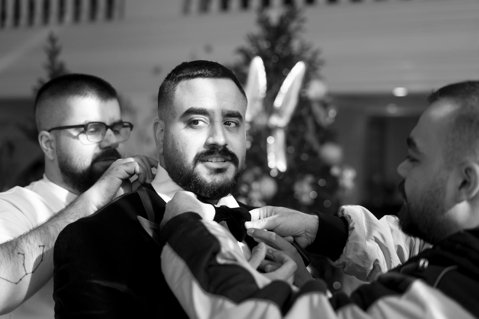 A groom is assisted by friends in adjusting his suit, inside a festive venue.