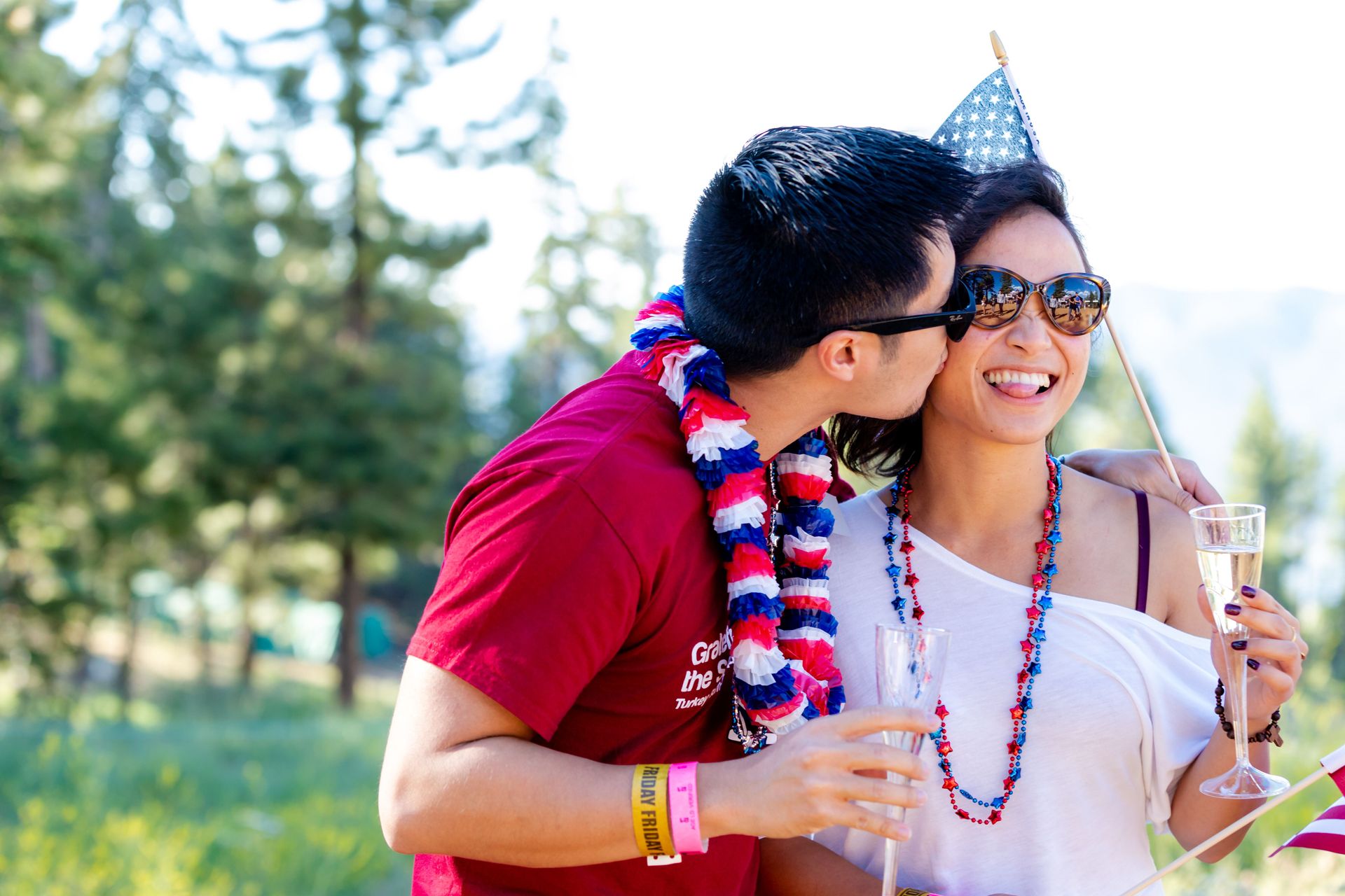 Couple celebrating outdoors with flags and drinks, embracing Fourth of July spirit.