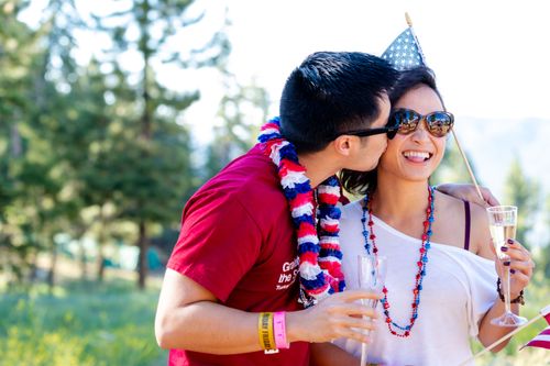 Free Couple celebrating outdoors with flags and drinks, embracing Fourth of July spirit. Stock Photo