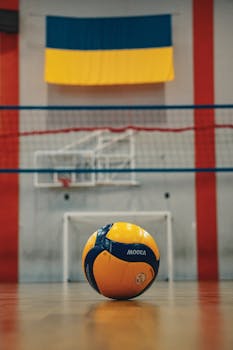 A volleyball rests on a gym floor in Kyiv, Ukraine, with the Ukrainian flag and net in the background.