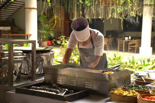 A chef in an open outdoor kitchen prepares dishes on a hot griddle surrounded by fresh ingredients.