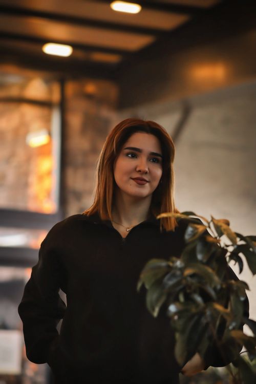 Free A young woman stands indoors near a plant, with warm ambient lighting. Stock Photo