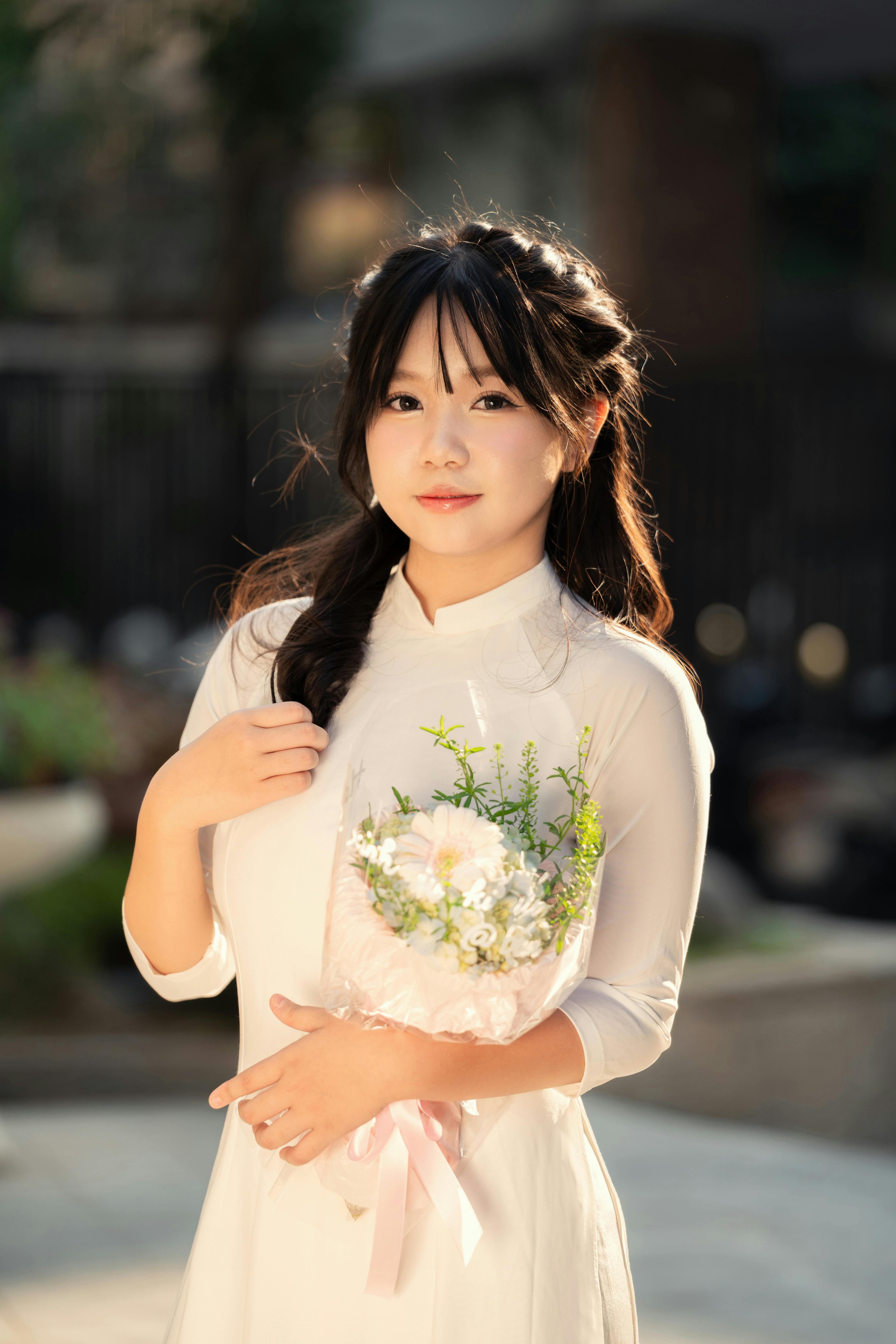 Elegant young woman in white áo dài holding flowers in a serene outdoor setting.
