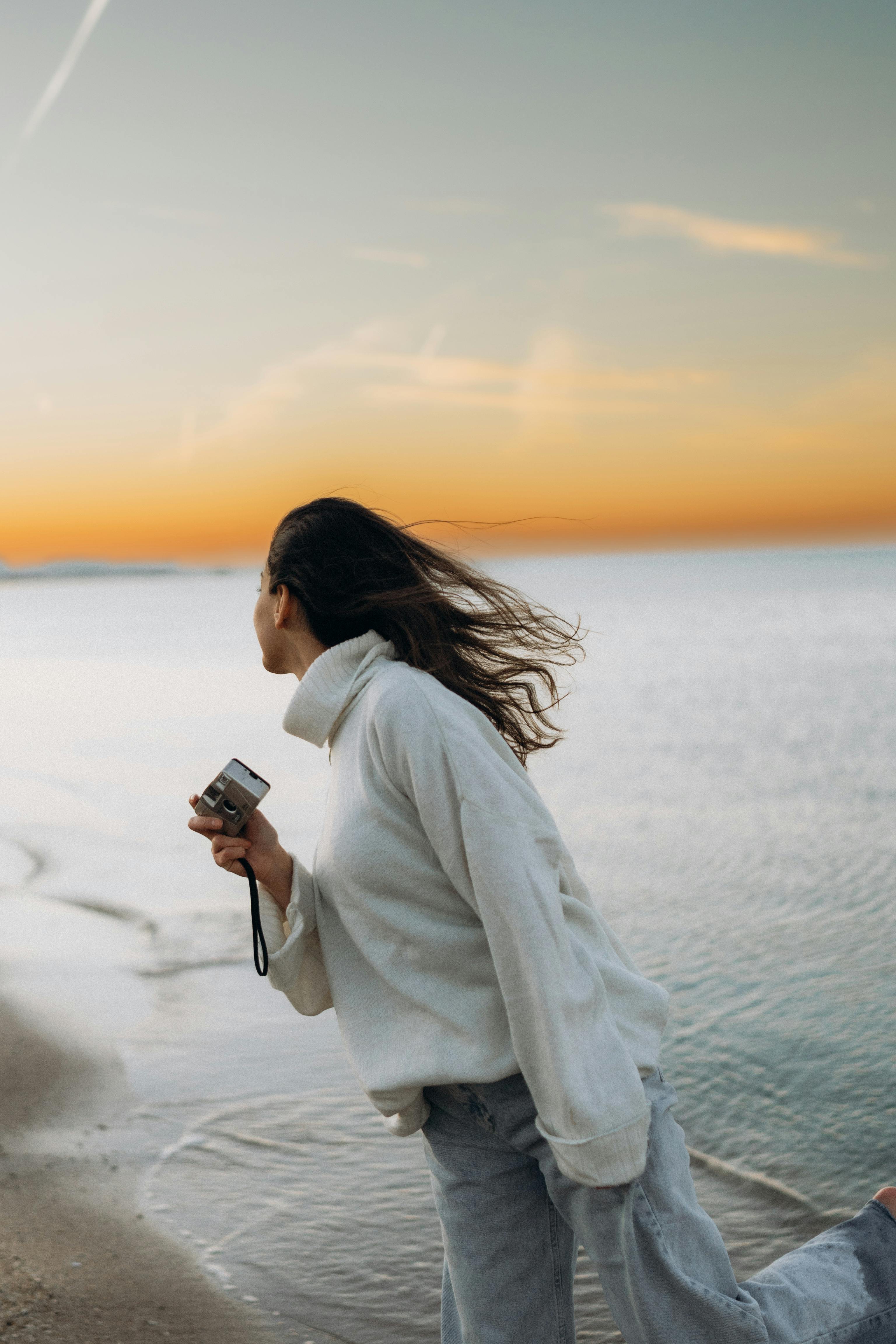 A woman with flowing hair holds a camera by the beach during a serene sunset.