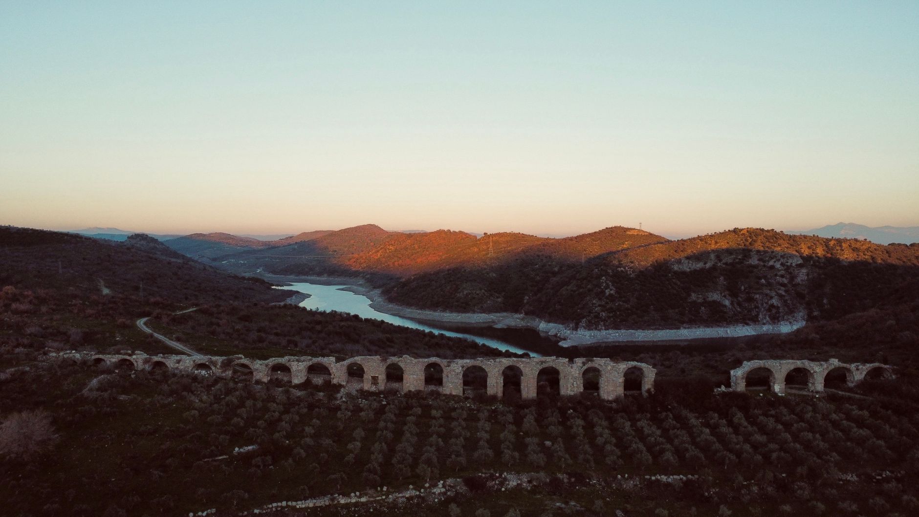 Aerial view of an ancient aqueduct crossing a scenic river valley during a peaceful sunset.
