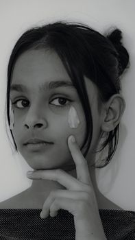 Black and white portrait of a young girl applying skincare cream with focus on eyes and thoughtful expression.