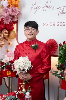 Young man in a red traditional outfit holding a bouquet at an indoor wedding ceremony.