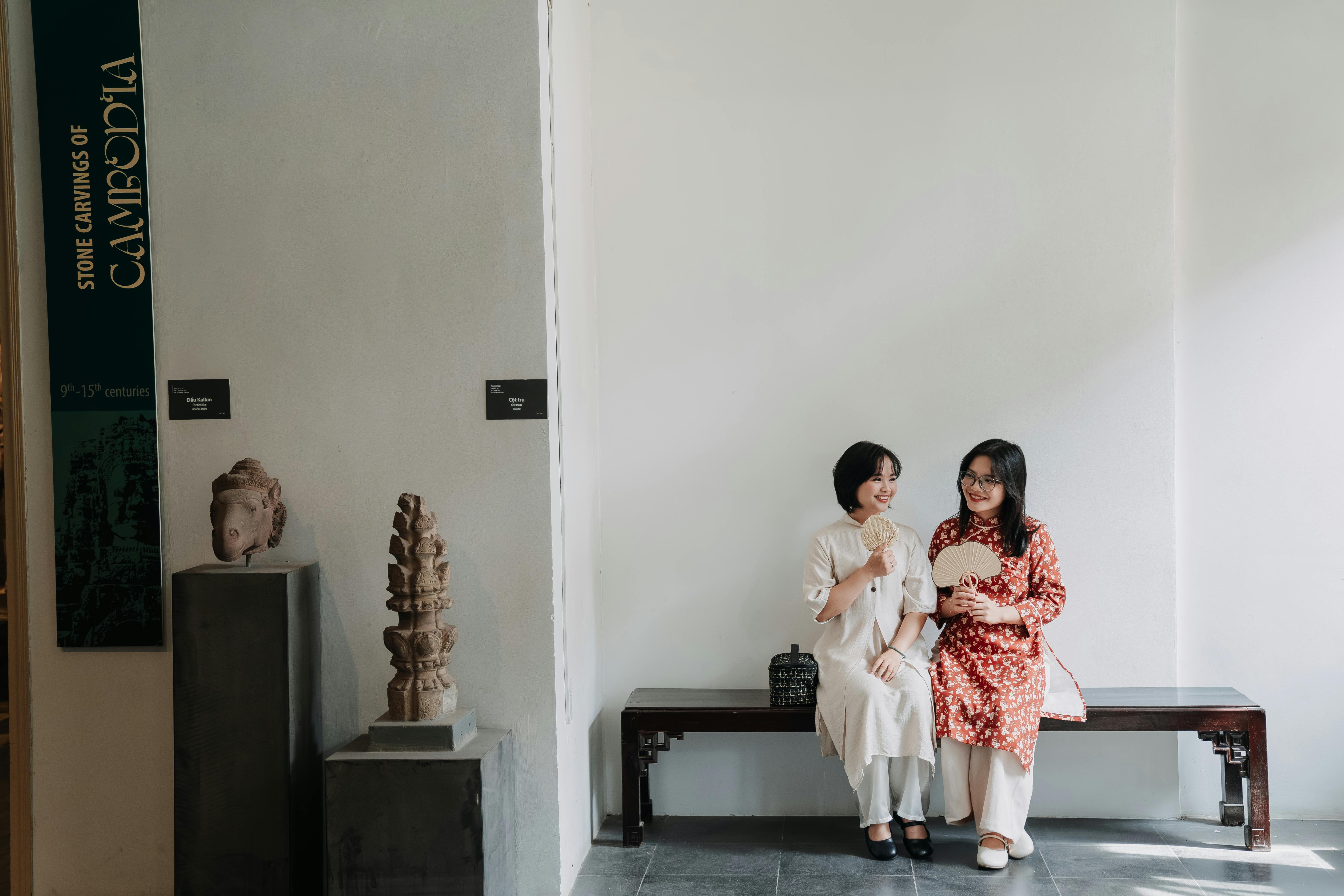 Two women sit on bench near Cambodian stone carvings in gallery.