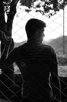 Black and white silhouette of a man standing behind a fence, capturing the essence of solitude at sunset.