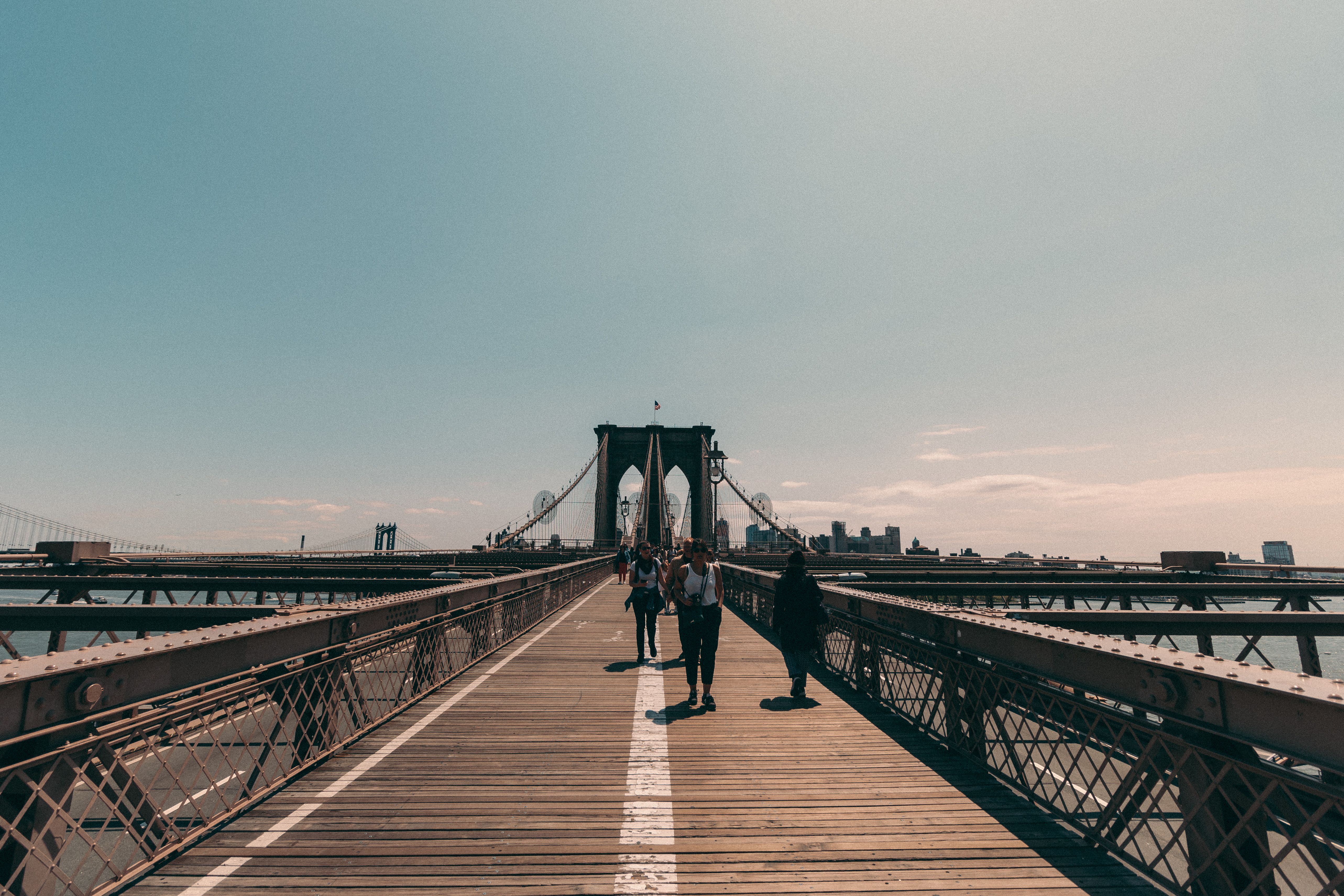 Brooklyn Bridge pedestrian path with tourists walking toward Manhattan skyline visible in the distance