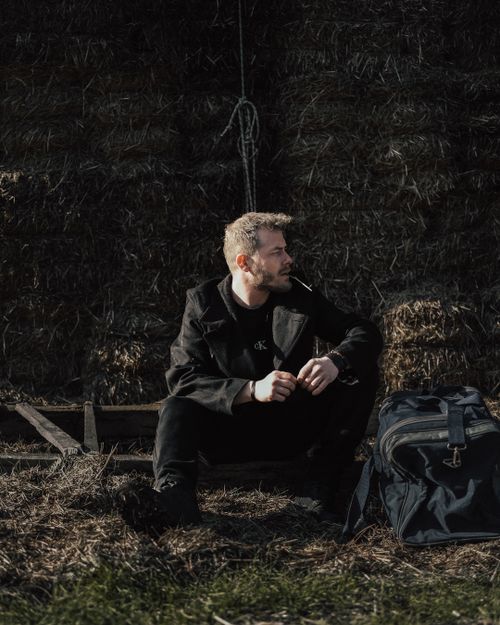 Free Man in black sits near hay bale in Türkiye, smoking with a backpack by his side. Stock Photo