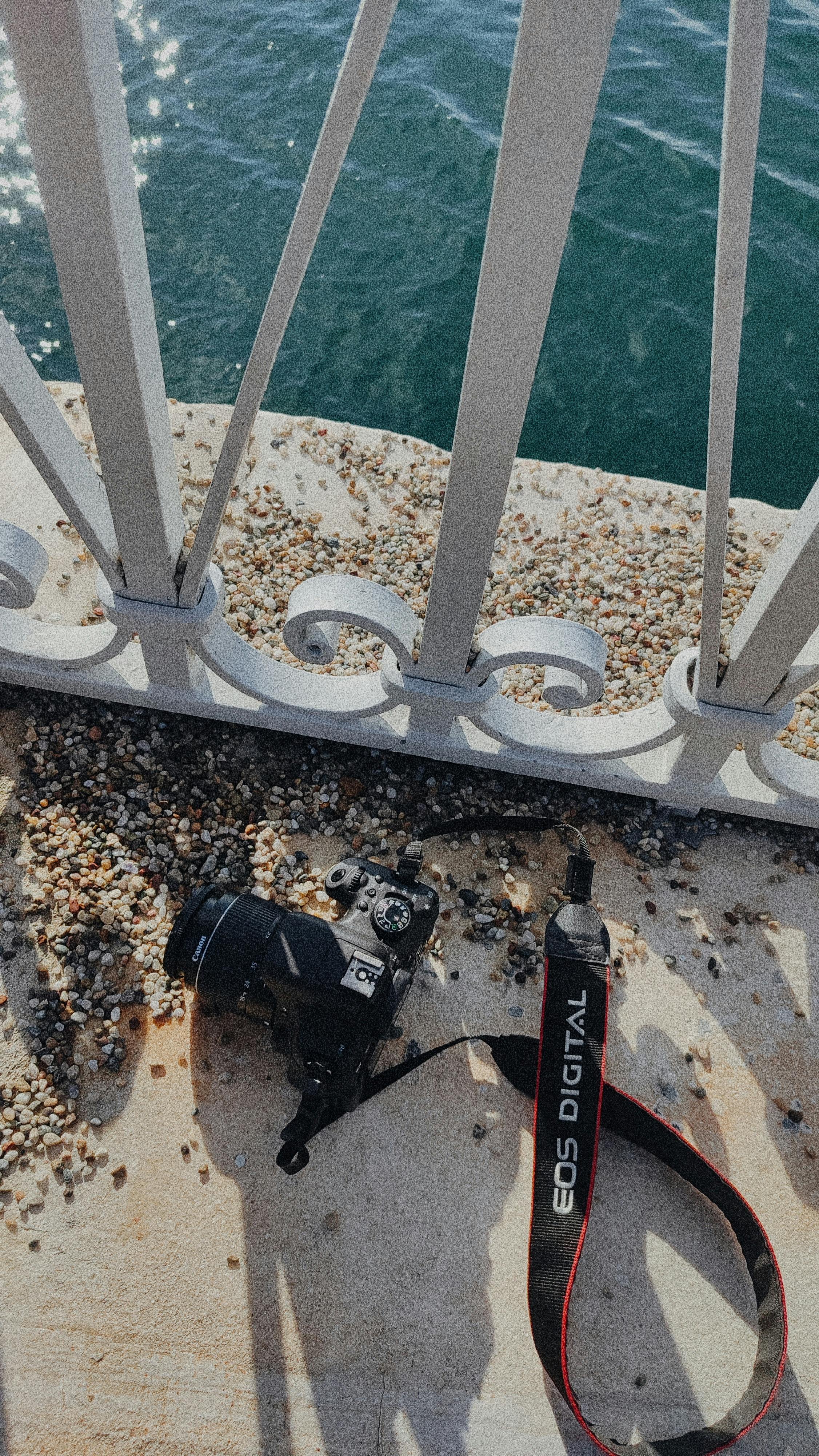 A camera rests on a pebbled surface by a waterfront, under a metal railing.
