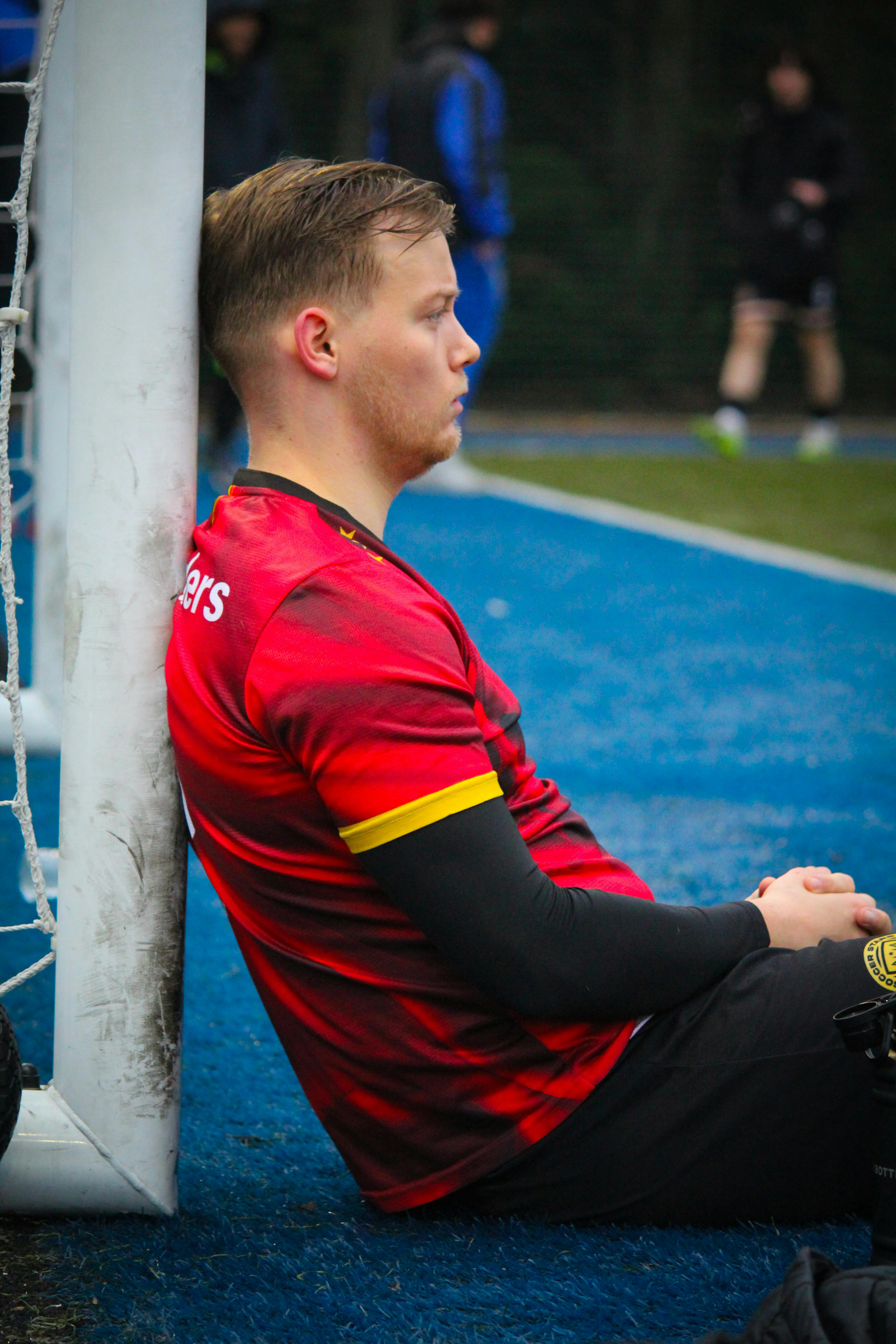 Soccer player in thoughtful pose during break sitting by the goal post on a field.