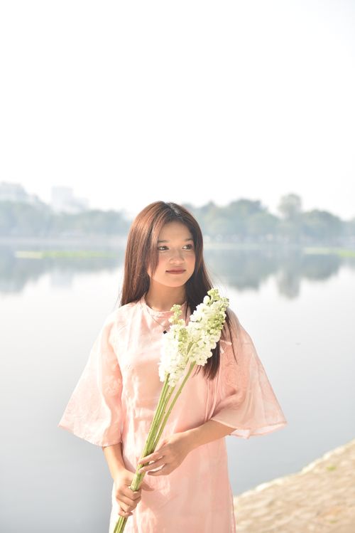 Free Elegant young woman in pink holding flowers by a serene lake on a sunny day. Stock Photo