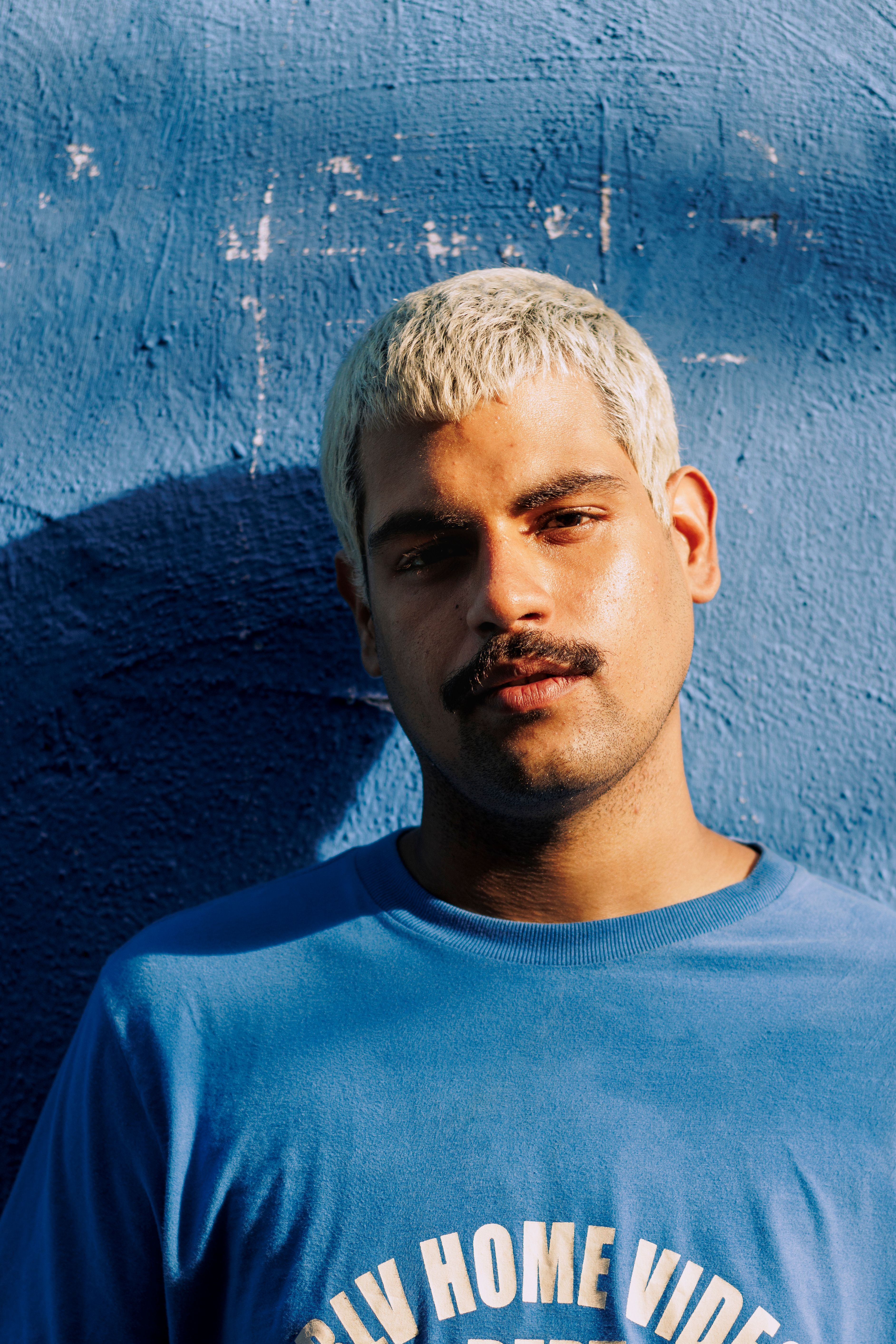 Man with platinum blonde hair and blue shirt standing against textured blue wall.