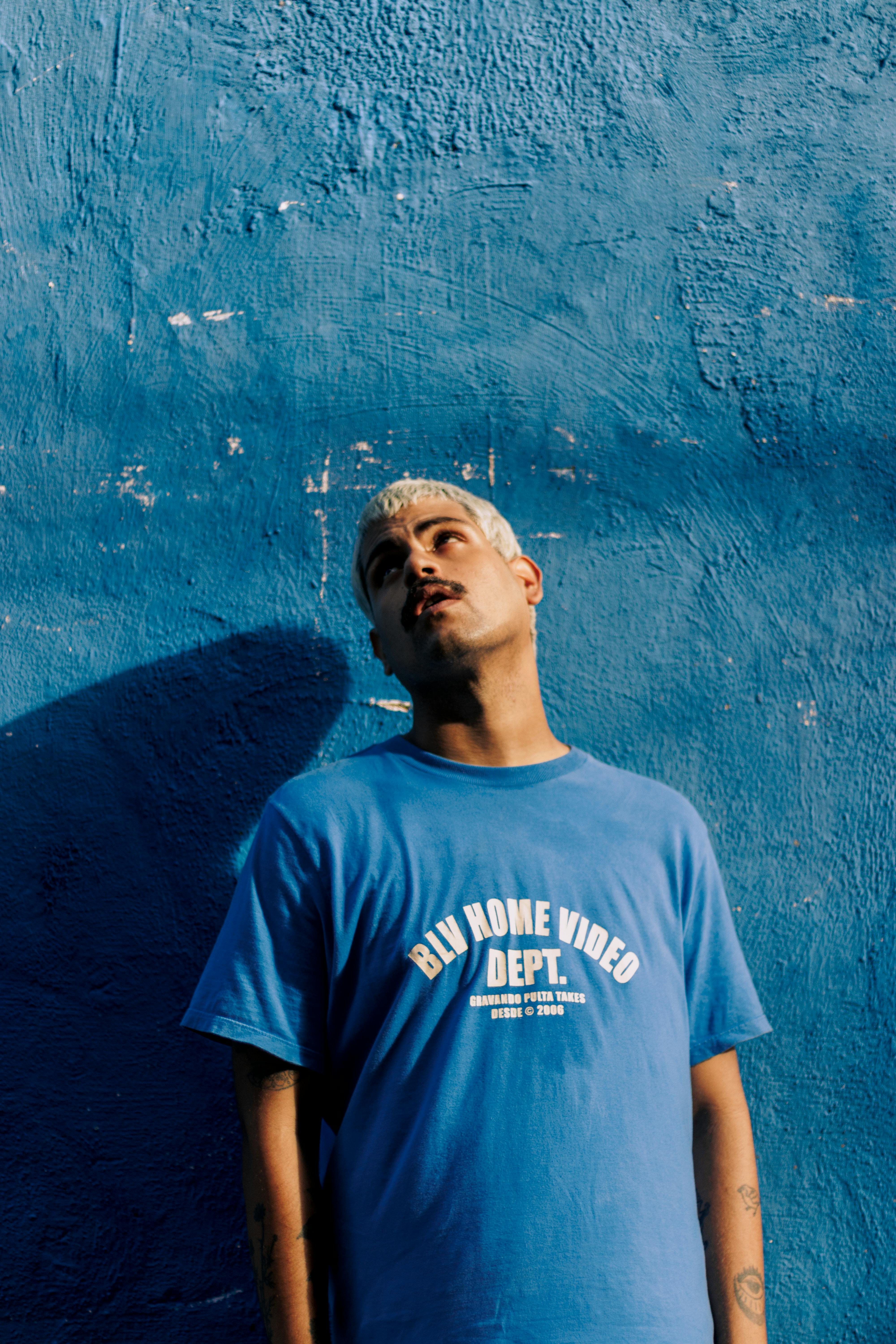 Young man poses against a textured blue wall in casual attire under bright daylight.