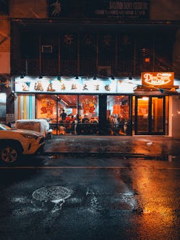 A bustling Asian restaurant glows warmly at night, reflecting on a rain-wet city street.