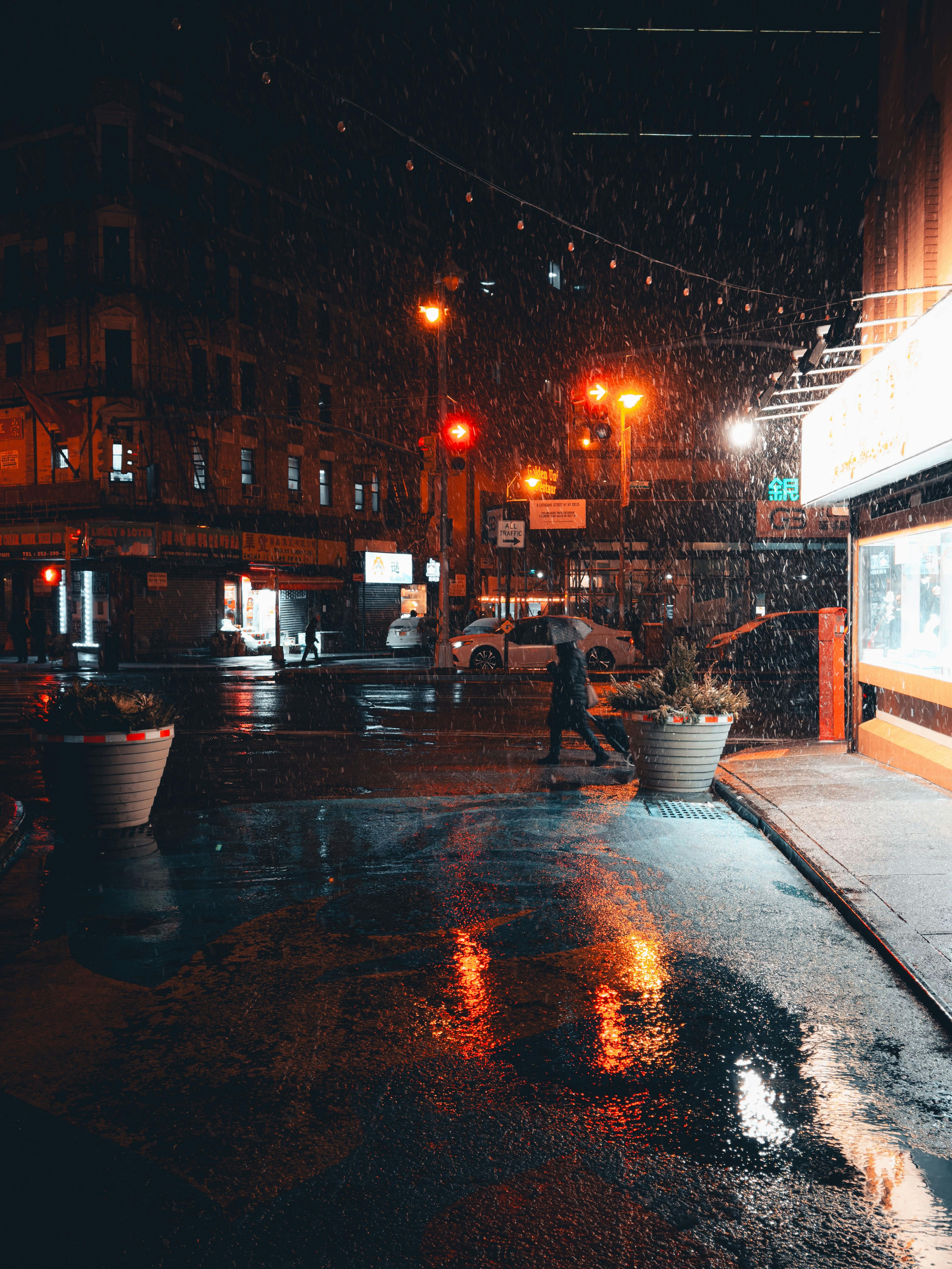 Nighttime urban scene with rain, reflections, and a lone pedestrian crossing a dimly lit street.