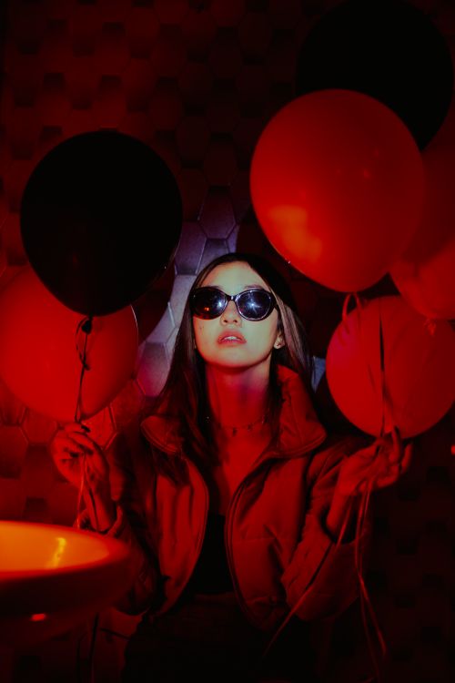 Free Chic woman in sunglasses holding red and black balloons in dimly lit room with moody ambiance. Stock Photo