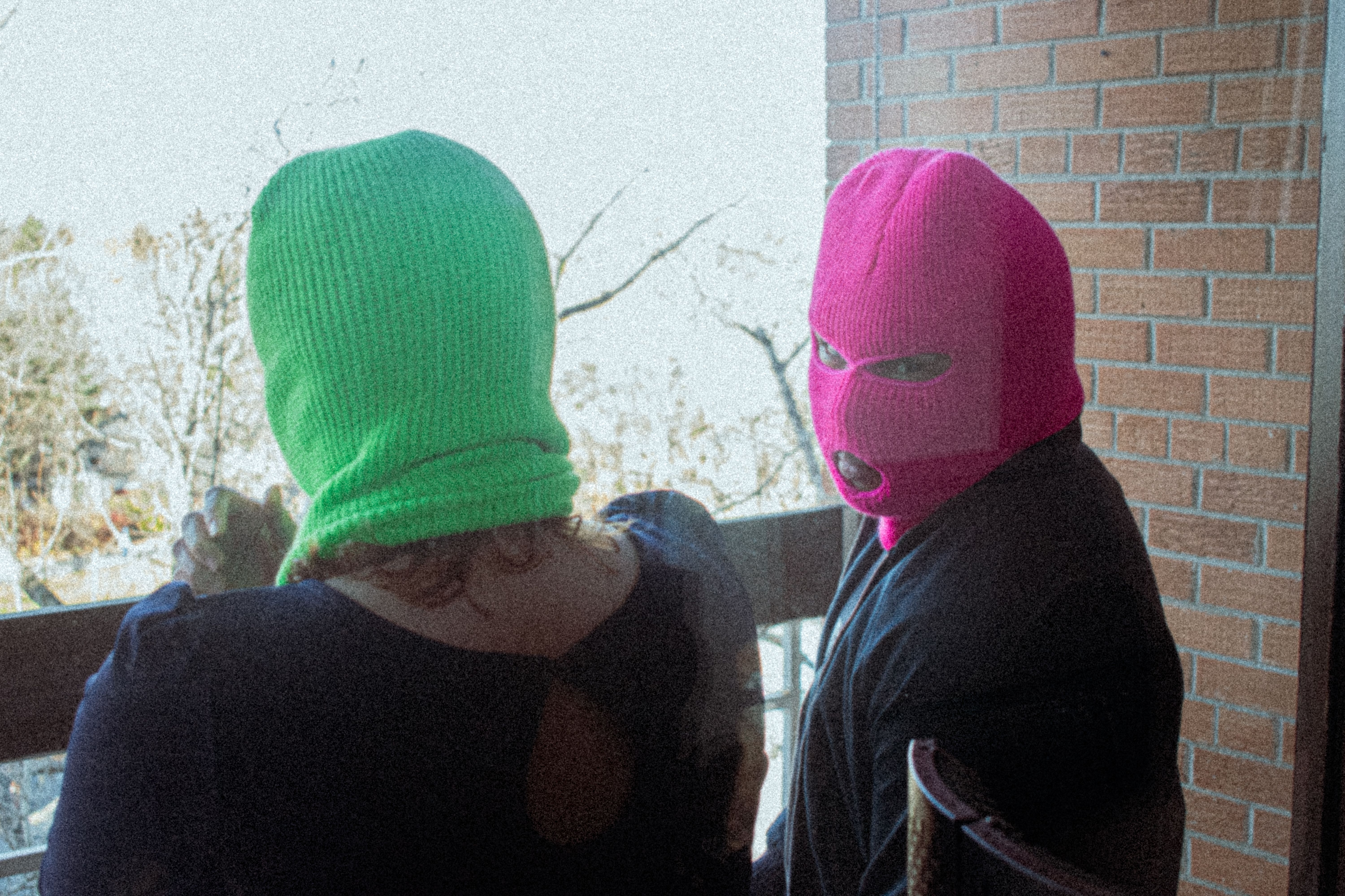 Two adults wearing neon ski masks on a balcony overlooking a winter landscape.