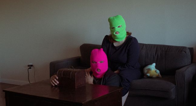 Two people wearing colorful ski masks sit indoors with a mysterious box.
