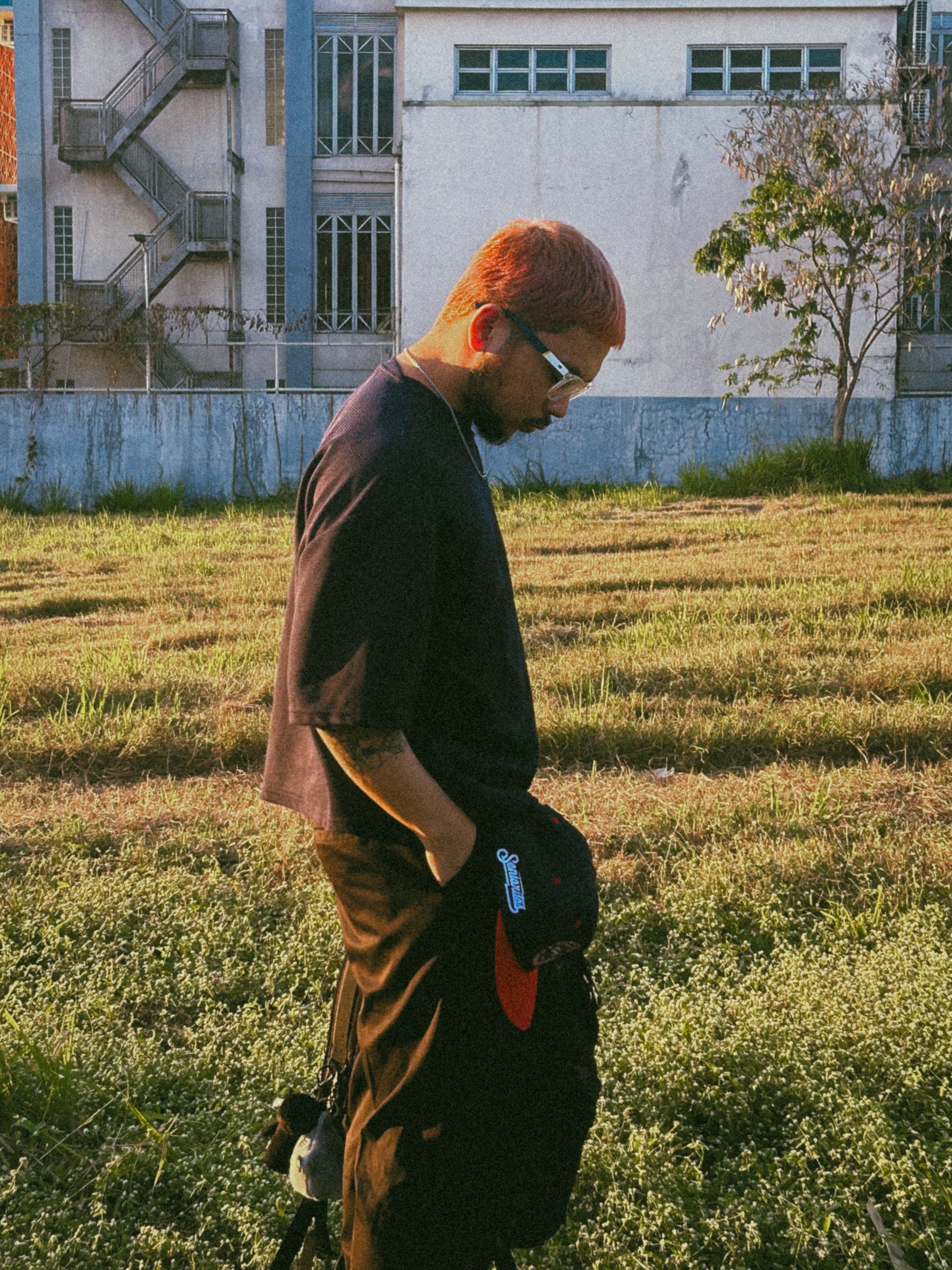 Man standing contemplatively in a grassy urban area with a background of buildings.