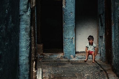 Free A young boy sits in a narrow, dimly lit alleyway with blue walls in Morocco. Stock Photo