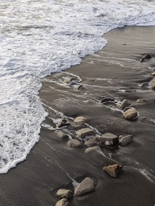 Free Gentle waves washing over a rocky shoreline with black sand, creating a serene coastal scene. Stock Photo