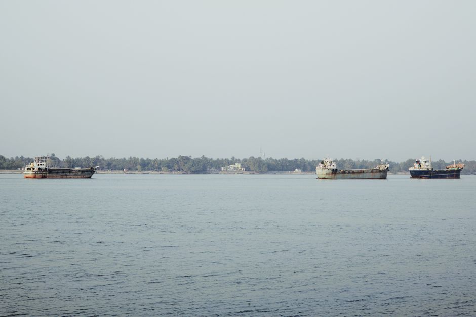Three cargo ships float peacefully on a serene river under a clear sky, with a lush forested shoreline.