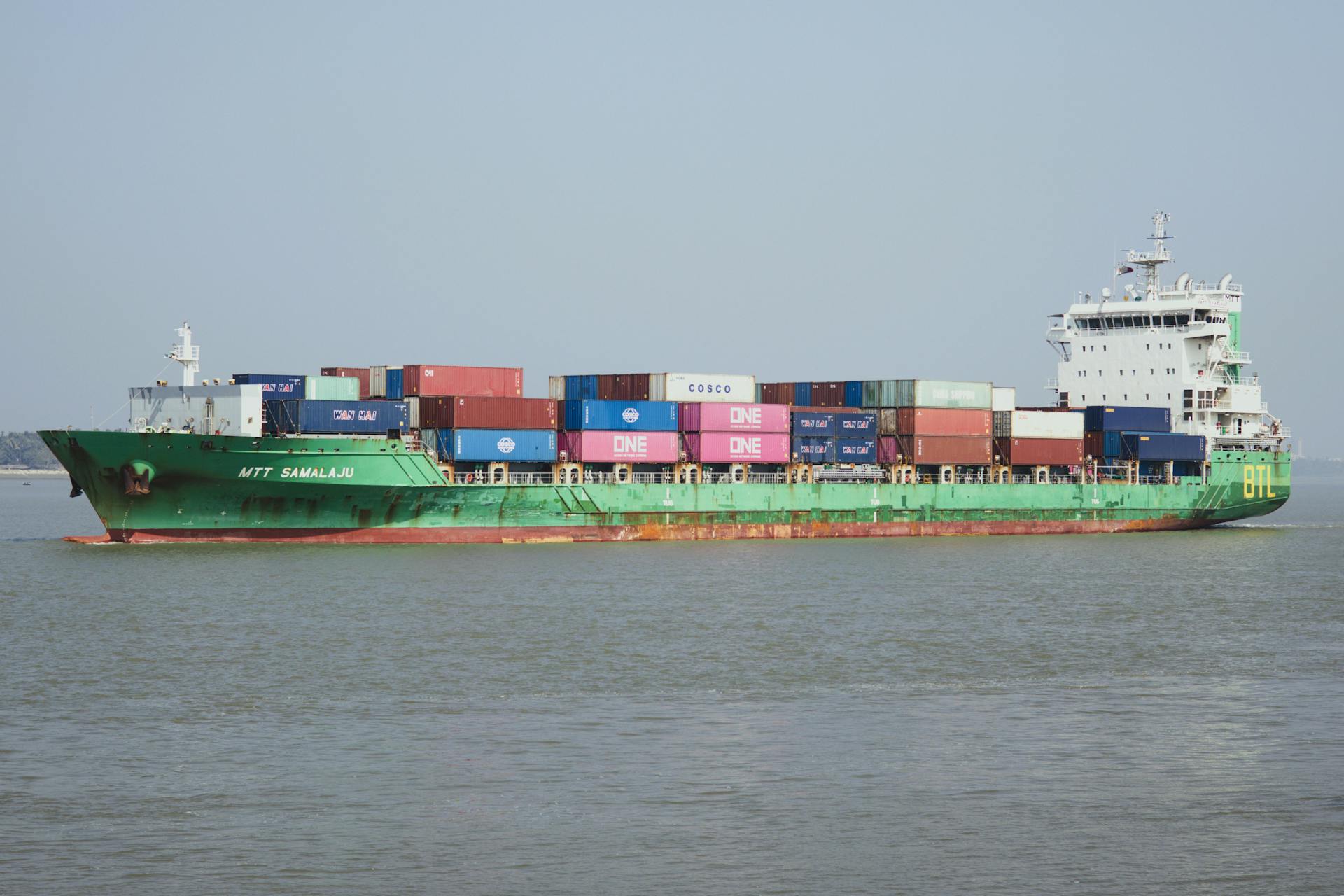 A cargo ship carrying multiple containers navigates through calm waters under a clear blue sky.