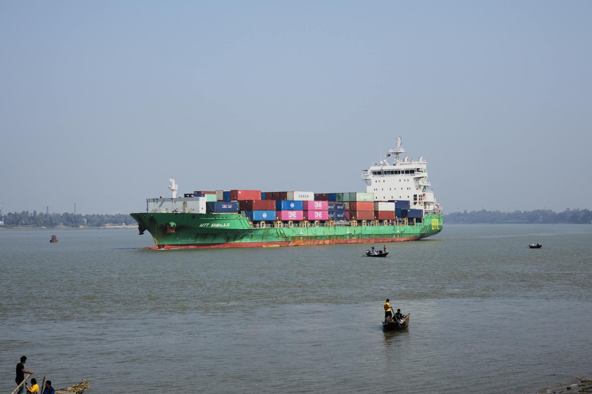 A colorful cargo ship carrying containers sails peacefully on a river, with small boats nearby.