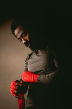 A focused male boxer wrapping his hands with red bandages in dim lighting.