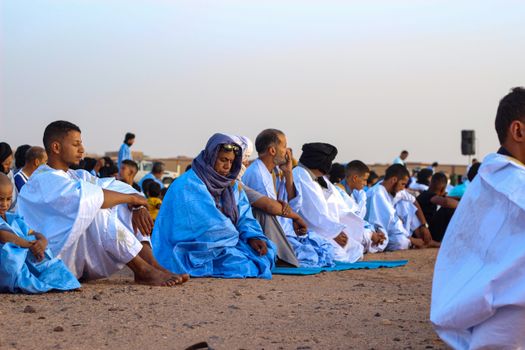 A group of people dressed in blue and white robes gathered for prayer in a desert landscape during daytime.
