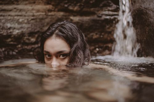 Free A woman partially submerged in a calm pool, near a waterfall, conveying tranquility. Stock Photo